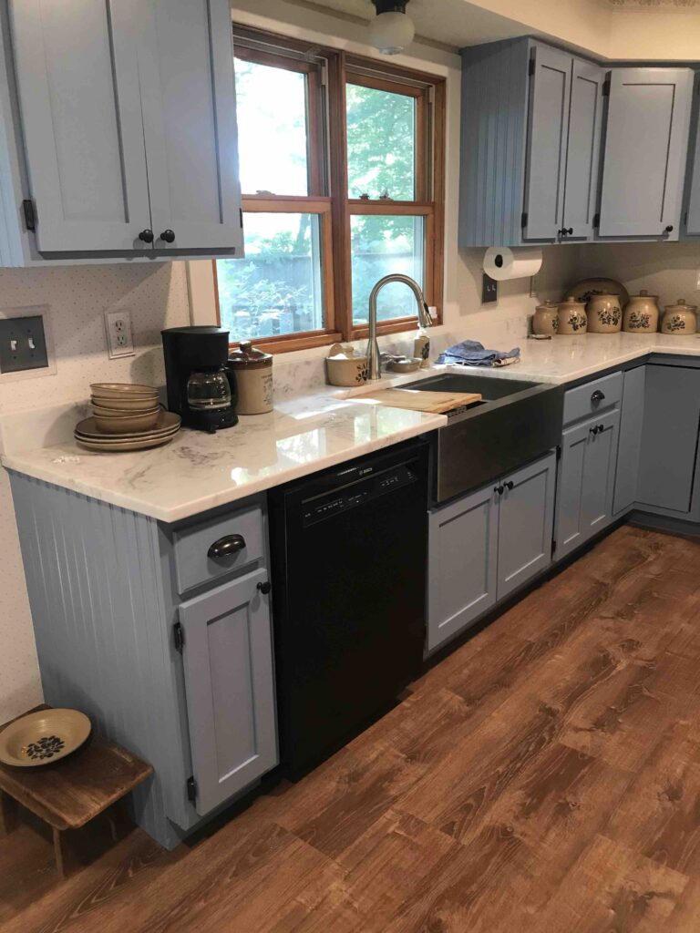 Kitchen renovated with grey-blue cabinets, white countertops, new hardware, farmhouse sink.